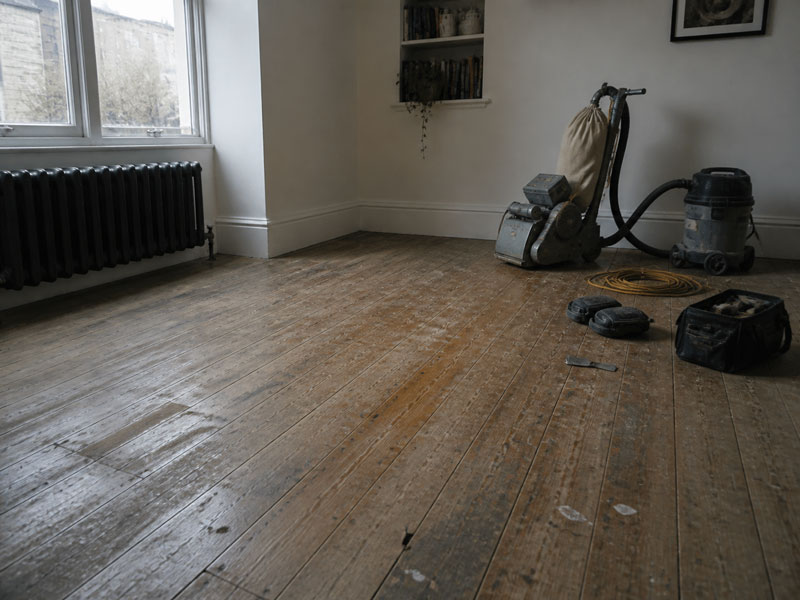 Bare, worn original pine floorboards in an empty room before sanding, with the sanding machine and dust extractor set up ready to start