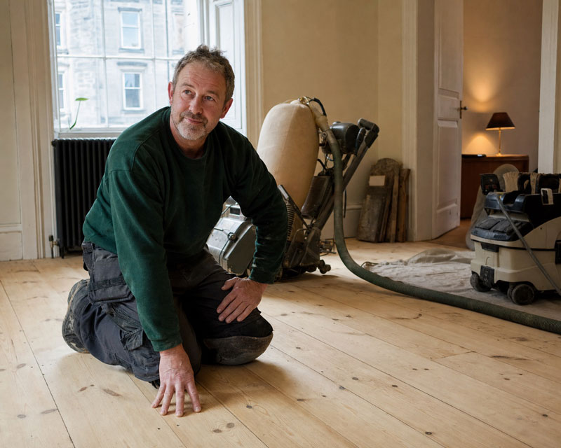 Ian Forsyth kneeling beside a freshly sanded pine floor mid-job, sanding machine and dust extractor visible behind him