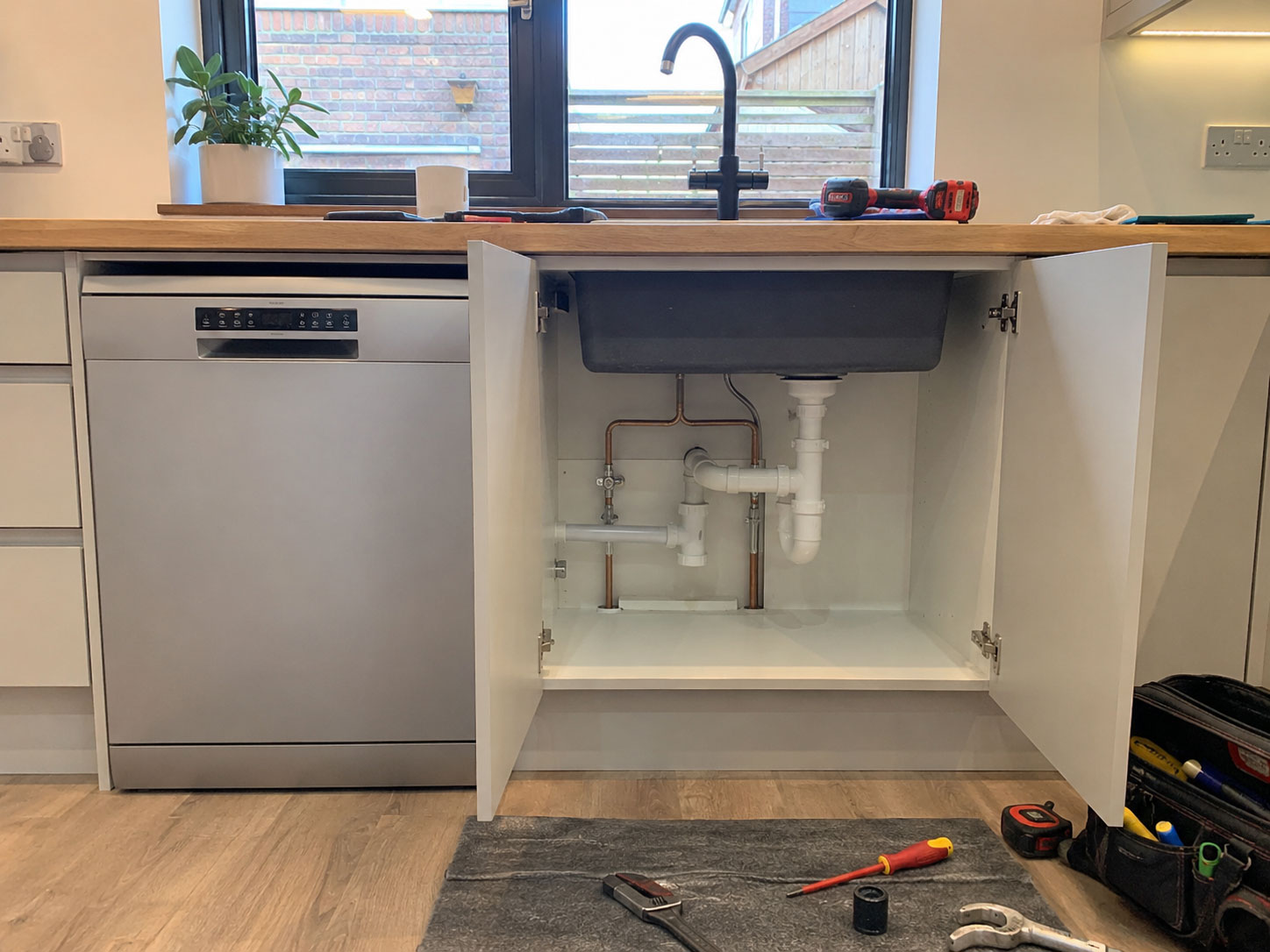 Open cabinet beneath a new kitchen sink showing freshly installed copper and waste pipework, with an integrated dishwasher connected alongside